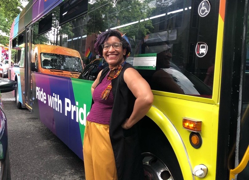 Kristen in the colors of the nonbinary flag leaning against a WMATA metrobus in the progress pride flag colors
