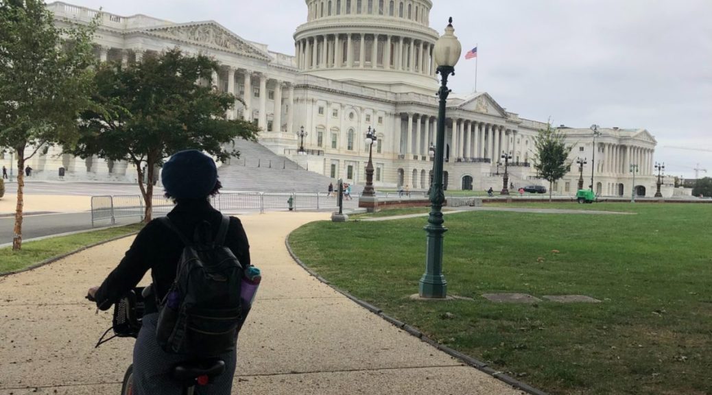 Kristen on a bike in a shadowy foreground looking up at the US Capitol on an overcast day