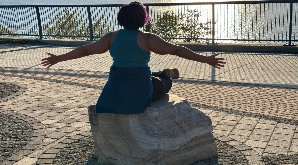 Kristen, arms outstreched, sitting on a rock under the sun facing the Potomac River