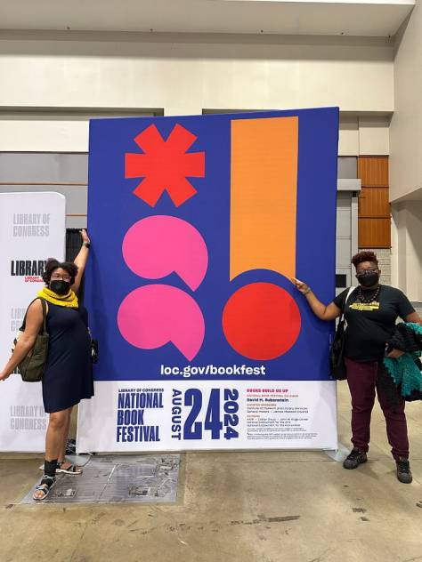 Kristen and Les standing and demonstrating the large poster for the Library of Congress National Book Festival in August of 2024 on the book signing and sales floor at the Washington Convention Center.