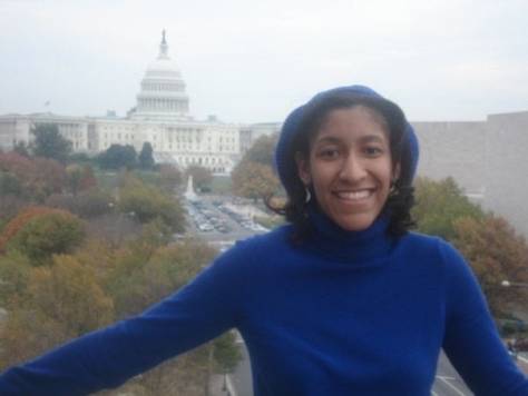 Kristen standing in a blue turtleneck and a blue sweater, with straightened but feathered Black hair on a balcony with the West front of the US Capitol building and a cloudy sky in the background.