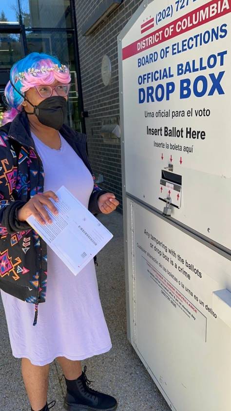 Kristen, in a wig that is reminisent of cotton candy, stands in front of a DC official ballot box.