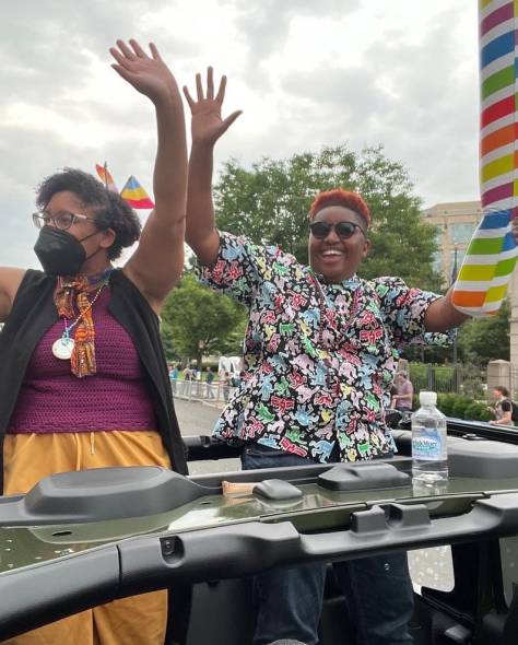 Kristen and Les waving to a crowd dressed in Pride clothing as they ride in the back of a Jeep.